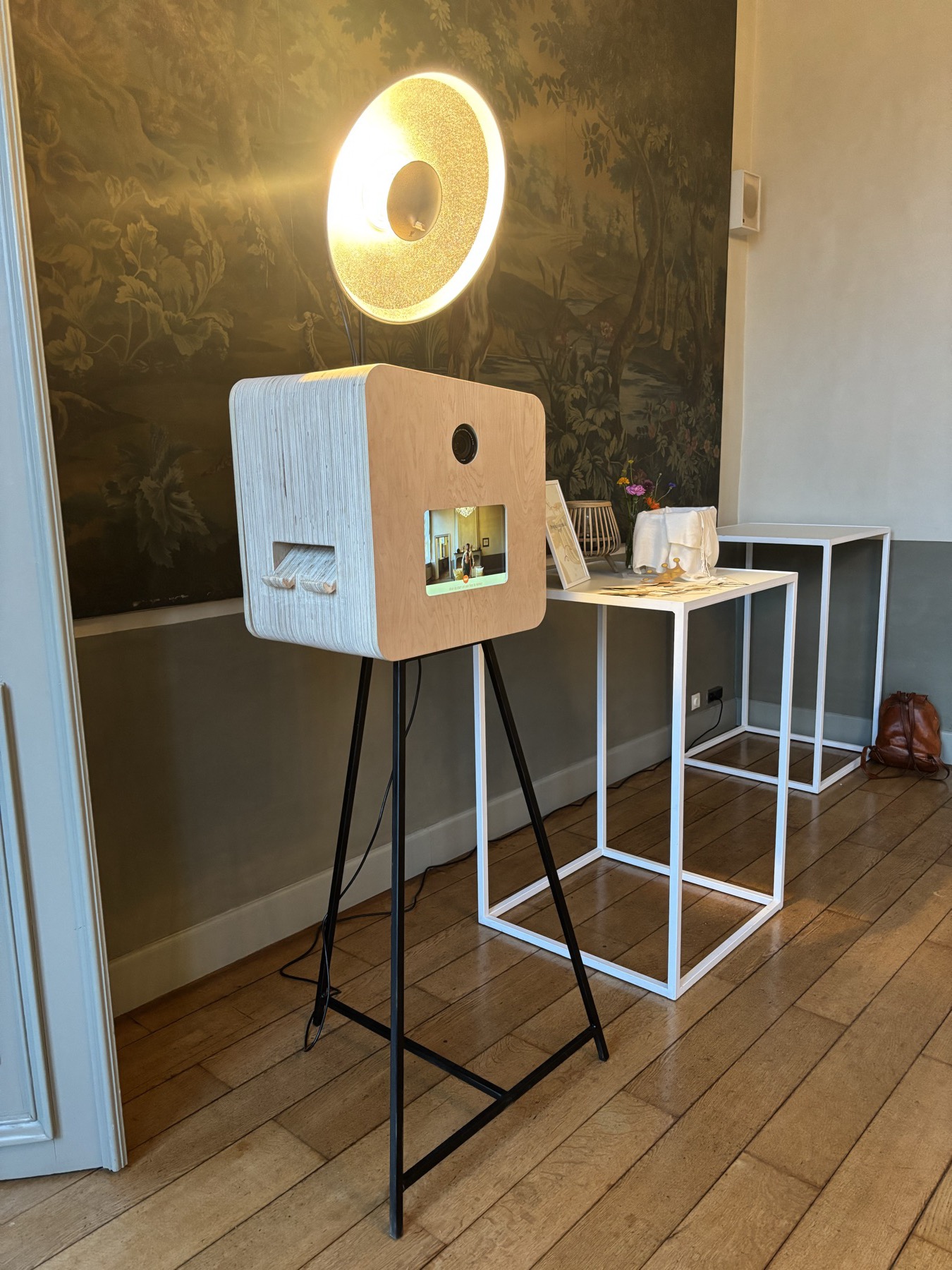 De houten photobooth op zijn zwarte statief, met de gouden lamp erboven, voor een geschilderd behang in een kasteelzaal